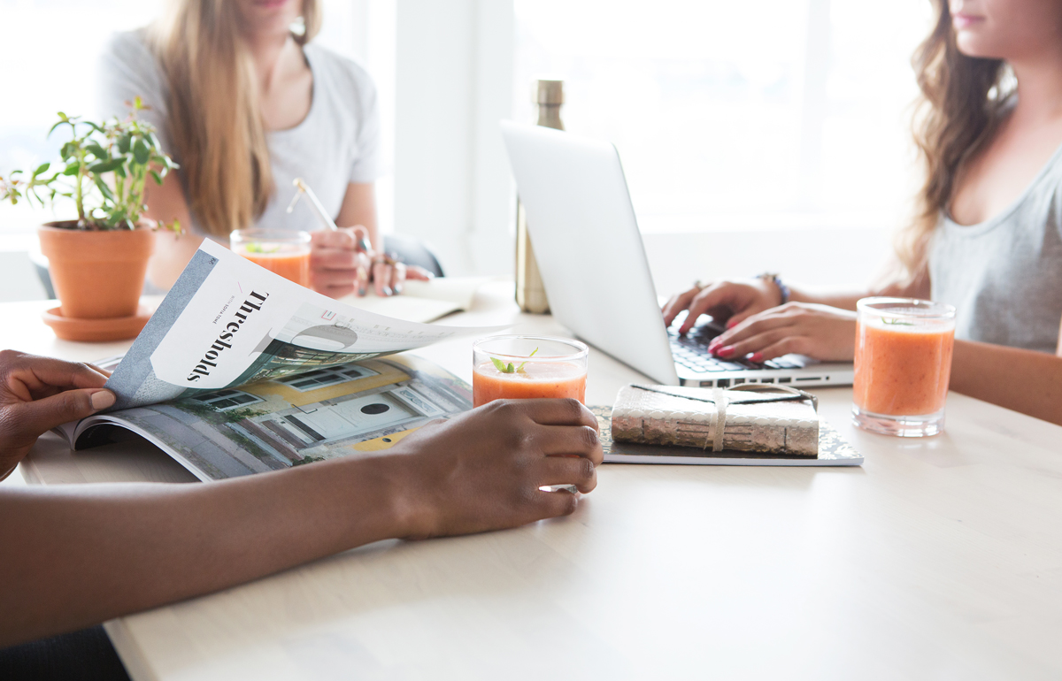 Drei Frauen sitzen am Tisch. Du siehst nur die Arme und keine Gesichter. Eine mit Laptop daneben ein Getränk eine schreibt, eine liest ein Magazin. Das Bild soll Freundschaft suggerieren.