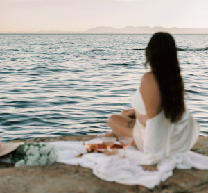 Frau mit dunklen, langen Haaren sitzt am Meer und genießt den Sonnenuntergang auf einer Piknickdecke. Sie trägt ein weisses Hemd die Schulter liegt Frau. Sie hat wunderschöne Kurven und ist mit sich zufrieden.