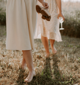 Zwei Frauen aber Mitt eKörper in beigen Kleidern. Eine trägt ihr Paar Schuhe die andere ein Weinglas mit Weißwein. Si gehen Barfuss über Gras.