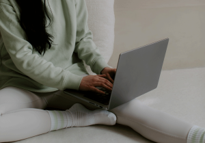 Frau mit dunkeln langen Haaren, sitzt auf dem Bett mit einem Laptop auf einem Bein. Sie schreibt etwas. Sie trägt ein Salbei grünes Oberteil und eine Beige Farben Leggings. Weisse hohe Socken. Das Bild wirkt entspannt.